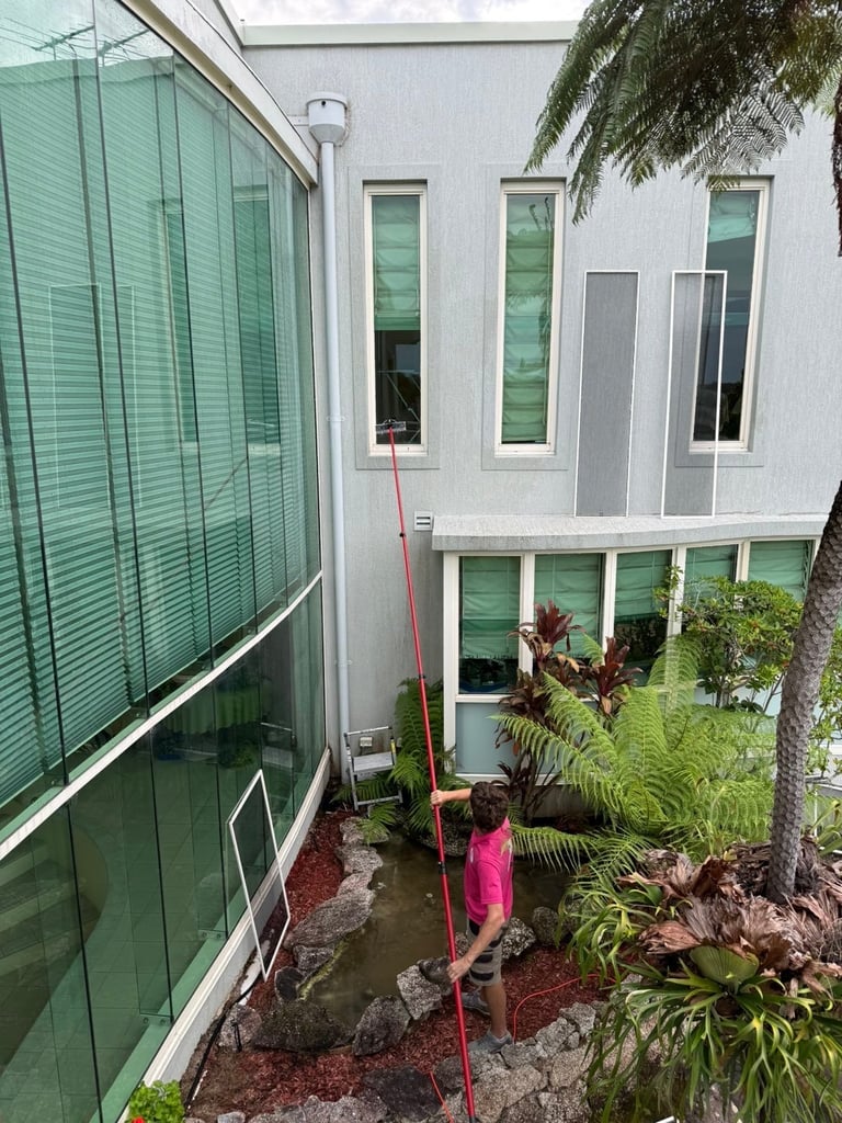 Person in pink shirt standing in narrow courtyard between white modern building and large green mesh water tank, surrounded by tropical plants